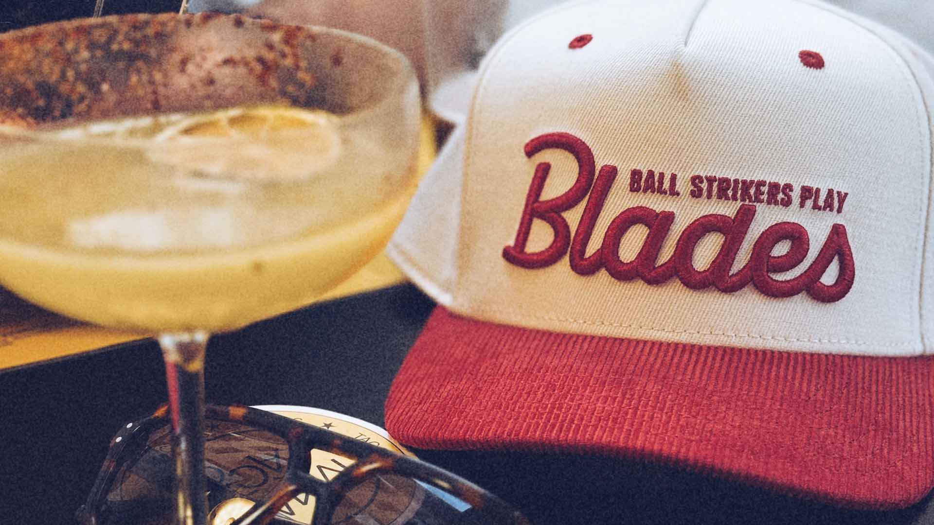 Cap with 'Ball Strikers Play Blades' text next to a cocktail glass on a dark surface